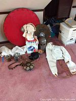 Overview of lot on pink carpet showing red bass wood shield, smaller metal shield, metal helm, white shirt and jacket, leather pouch, canteen, coifs, mugs, bowl and necklaces.
