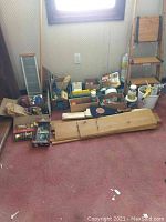 Wide view of the lot showing wooden planks, boxes of hardware, three-step wooden ladder, buckets with painting supplies and tools on carpeted floor near window.