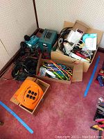 Overview of lot of electrical supplies including extension cords coiled on floor, boxes with various cables, cord ties, surge protectors, and electronic devices