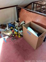 Photo showing pile of assorted leathercraft items, large piece of brown leather and cardboard box on pink carpet.