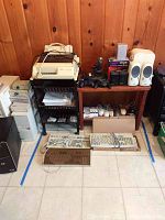 Wide view showing lot of computer accessories including printer on rolling cart, four keyboards on floor, white computer mice, joysticks, speakers on wood table.