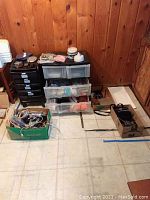 Overview of lot showing two plastic storage units with multiple drawers and wheels, boxes with assorted wires and components on floor against wood-paneled wall.