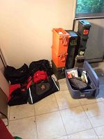 Nine ammo crates stacked side by side against wall, including orange plastic crates and metal crates with insignias, adjacent to black and red shoulder bags and gray plastic container with firearm accessories.