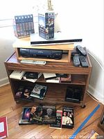 Media cabinet containing black Panasonic VHS player, Sony DVD player, remotes on top shelf, VHS and DVD box sets on lower shelves.