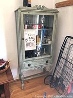 Front view of the vintage cabinet showing glass door with display contents inside, carved top and apron details, and single drawer.