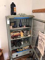 Full view of wooden cabinet with glass door open, showing various items inside including bud vases, candles, oil lamp, and safety light sticks.