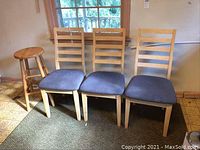 Three wooden ladder-back dining chairs with blue fabric seat covers and matching wooden round stool grouped together.
