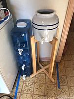 Photo showing wooden water cooler stand with ceramic dispenser on top and three blue plastic water dispensers stacked beside it.
