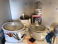 Lot includes two crockpots, one with vegetable design and glass lid, two Juiceman juicers, and a boxed KitchenAid food processor.
