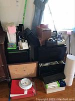 Photo showing assorted office supplies including stapler, shredder, plastic organizers, hanging folders, corkboard, wall clock, push pins, copy paper, and rolling file cabinet.