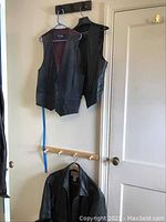 Photo shows two black leather vests hanging on wall hooks and a black leather jacket hanging below them, with a pair of black leather boots on the floor below the jacket.