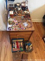 Photo shows wooden dresser with assorted costume jewelry spread out including earrings, brooches, bracelets, necklaces, watches, cufflinks, and rings. Also visible are two wooden carved boxes on the floor and two jewelry boxes containing additional pieces.