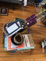 Overview of the lot with stacked items including baskets, curtain panels in packaging, drapery rods, placemats, dish saver kit, and a wine box.