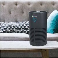 GermGuardian air purifier placed on a table in a living room setting, showing its black cylindrical shape with air vents and a blue circular button on front.
