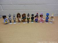 Front view of 11 Wizard of Oz mini dolls standing side by side on a wooden surface against a white-brick wall, each with individual white plastic stands.