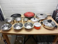 Photo showing the assortment of pots, pans and mixing bowls arranged on a table with white brick background.