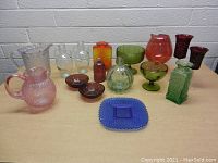Overview photo of 15 colorful glass items on table against white brick wall, showing variety of colors and shapes including pitchers, bowls, vases, glasses, and dishes.