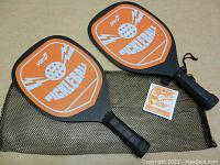 Two new SportCo pickleball paddles with orange pickeball graphic face, black wrapped handles, original tag attached, and black mesh carry bag beneath them.