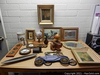 Wide view of lot on table with framed paintings at back, globe, wooden sculptures, plates, and model car in front.