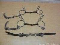 Top view of two vintage metal horse bits with attached curb chains and a leather strap with chain detail and buckles laid on a wooden surface.