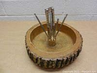 Round carved wooden nut bowl with bark edge, displaying the metal nutcracker and six metal nut picks upright in center wooden holder.