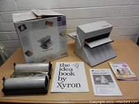 Photo of Xyron 850 laminator with two laminating rolls, manuals, idea book and original box on table against a wall.