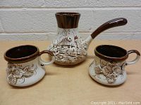Front view of set with teapot and two mugs showing details of leaf and flower textured design and dark brown glazing on rims and handles.