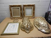 Two vintage gold ornate picture frames, a smaller framed artwork, and three decorative gold-toned wall plaques arranged on a table.