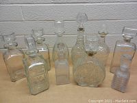 Photo showing 12 assorted clear glass decanters with lids, arranged in rows on a surface against a light wall background.