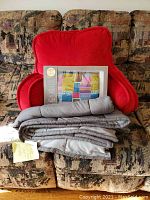 Overview showing red cushioned back rest chair, folded gray comforters, and boxed warming pillow on patterned sofa.