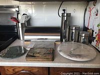 Kitchen countertop with stainless steel bread box, two marble hot plates, cork hot plates, white marble lazy susan, stainless steel storage containers, and utensil holder with serving spoons.