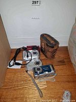 Four digital cameras on wood floor with charging cables and vintage leather binocular case.