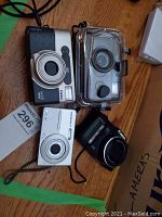 Four cameras placed on a wooden surface including two compact digital cameras, one film camera, and one camera in waterproof housing.