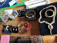Overview of assorted costume jewelry pieces laid out on a wooden table including snake design necklace and bracelet, earrings, rings, a beaded bracelet, and a decorative bowl and box.