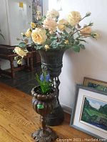Front view showing both metal standing plant pots with faux flowers inside, larger pot behind smaller pot, and a framed artwork leaning against the wall.
