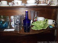 Wooden shelf displaying an assortment of china tea cups, saucers, ceramic bowl, clear glass mug with emblem, cobalt blue decorative bottle, and light green ceramic cabbage gravy boat.