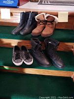 Four pairs of ladies shoes displayed on green carpet steps: two pairs of leather boots, one pair of brown shearling winter boots, and black and white knit running shoes.