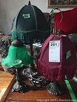 Photo showing four lamps: two with deep red fabric shades with tassels, one with dark green fabric shade with gold braid trim and tassels, and one with green frosted glass shade. Visible cords and ornate metal bases.