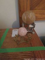 Two mineral globes on stands on wooden surface against wall background. Larger globe is yellowish-beige on ornate silver metal stand, smaller globe is pink on bronze-tone stand with dragon figures.