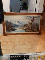 Front view of one original oil painting on canvas signed by Mitchell showing mountain, trees, lake, in a carved wooden frame, sitting on tiled floor