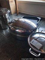 Image showing a large stainless steel roaster with lid, black grill pan, and stock pot lid on tiled floor near window.