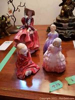 Five ceramic Royal Doulton figurines on table, including large lady in red dress and wide-brimmed hat, and four smaller girls in different colored dresses.