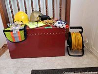 Photo showing red metal storage chest, two hard hats (yellow and gold), safety vests, protective eyewear, work gloves, and yellow extension cord on reel.