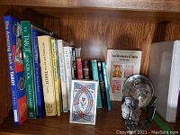 Shelf showing tarot books with titles including 'The Amazing Book of Tarot', 'The Tarot Handbook', 'Pictorial Key to the Tarot' and more, with tarot cards deck and glass globe on stand on the right.