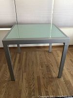Front view of square tempered glass-top dining table with brushed metal legs.