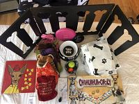 Black wooden dog gate with bone cutouts, multiple dog coats, Chihuahua-opoly game, original signed 'Weevo' dog painting on wood, skull dog dish, and various small dog accessories including tennis balls, lights, and potty pads.