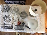 Overhead view of black metal kitchen decor arranged on white cloth, showing wire trivets, bottle stands, fruit baskets, decorative balls, and desk lamps.