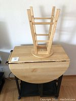 Wooden stool placed upside down on top of the table showing the stool's frame and legs, table with drop leaf extension down and two drawers visible in black metal frame.