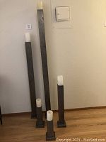 Five tiered rusted metal floor candle stands with white electric candles on top, arranged from tallest to shortest against a wall on wooden floor.