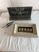 Photo of three items on white cloth: negative glass plate artwork on wood stand, 1957 Studebaker hood ornament, and framed set of five gargoyle heads.